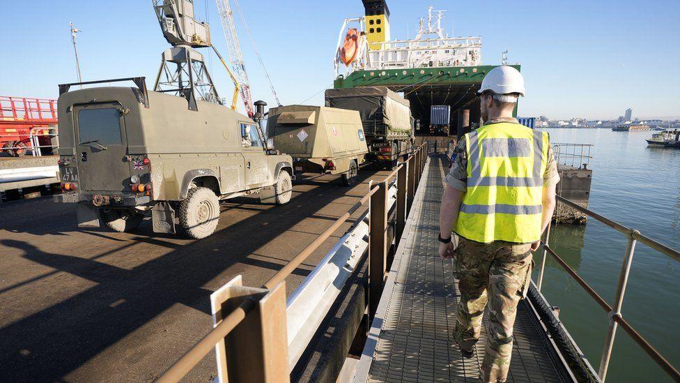 Man walking alongside army vehicles as they are driven onto a cargo ship - water and dock area can be seem to his right.