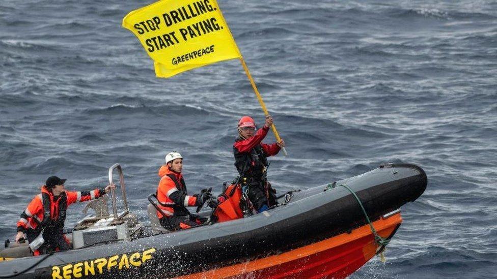 Three Greenpeace activists standing on an inflatable boat which is on the sea. One is at the wheel of the boat and a man at the front is holding a yellow banner that reads Stop Drilling Start Paying - Greenpeace. They are all wearing orange, red and blue waterproofs as well as helmets and baseball caps.