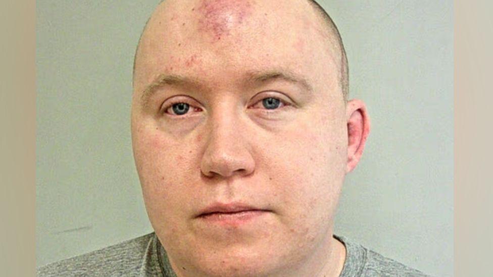Jacob Hill's mugshot. He is a man with receding, cropped light brown hair. He is photographed in a grey t-shirt.