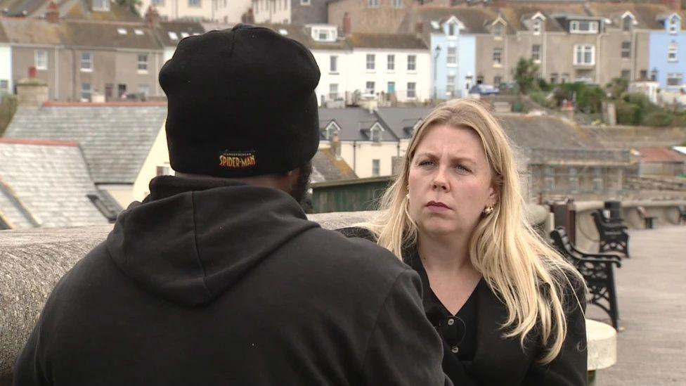 Cedrick, with his back to the camera, sat on a bench, as Sophie, who has blonde, shoulder length hair, is listening to his answer. Houses in Portland are behind them