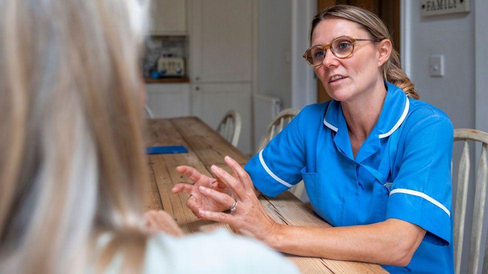 Over the shoulder view of an on call nurse talking to a patient who she is visiting at home. They are having a discussion about the patient's health.
