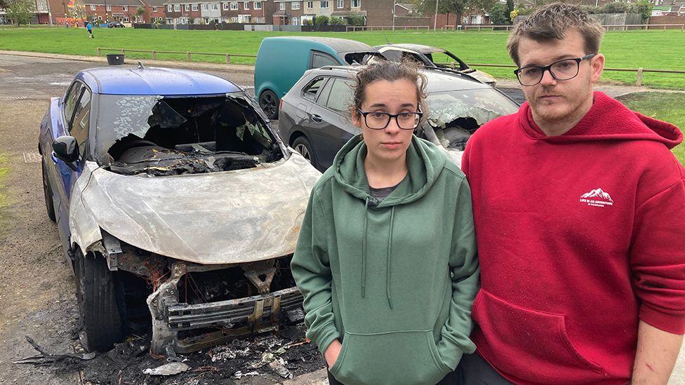 Shannon Rex and her partner Luke Deekens stand in front of their two burnt out cars. She has long dark hair pinned back behind her head and wears glasses and a green hooded top. He has short dark hair and wears glasses and a red hooded top.