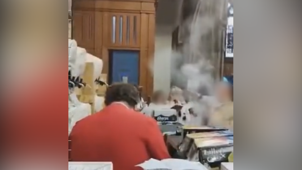 A blurry shot of the inside of Bangor Cathedral with multiple boxes and kegs of beers lining tables. A man with a red long-sleeved top on and dark brown hair is sitting with his back to the camera, other people are in the room but their faces have been blurred by the BBC for anonymity reasons. A blue door can be seen in the background underneath what appears to be a pulpit
