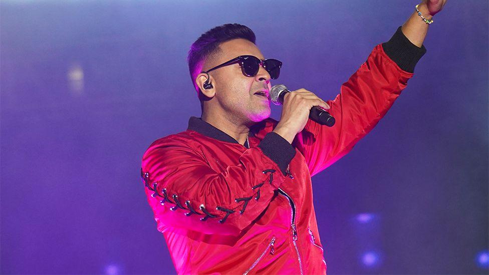 Jay Sean, a man holding a microphone on stage, wearing a red fleece and sunglasses.