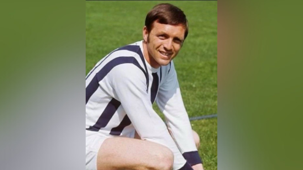 A young Jeff Astle looking directly at the camera and smiling, he is crouched down on the ground and dressed in his football kit.  