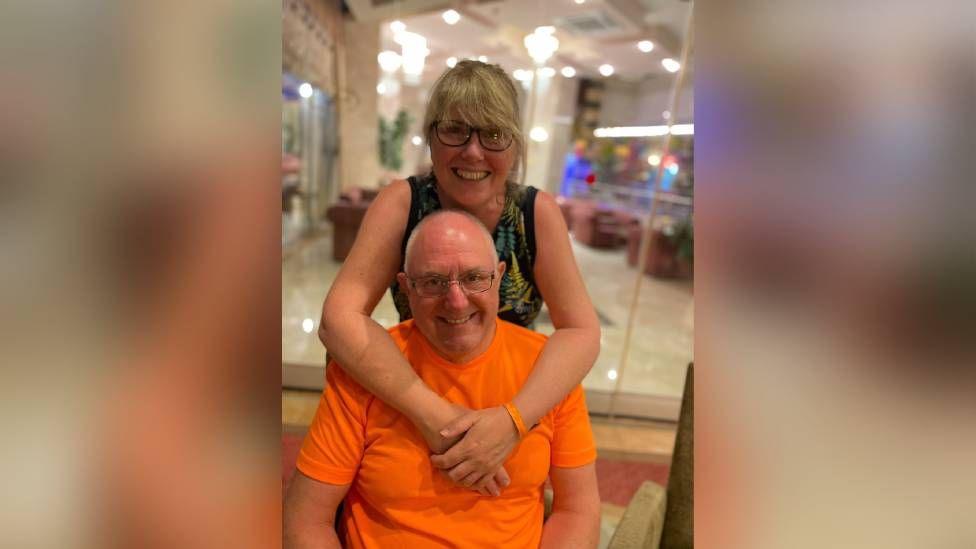 A couple with a woman with a blonde fringe and glasses stood behind a bald man with thin glasses wearing a bright orange shirt.