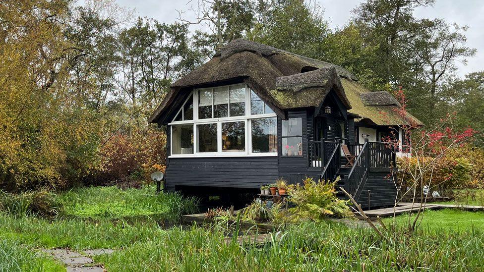 A black timber building with a mossy thatched roof and white windows. The building is built over a river. It is surrounded by grass with trees in the background 