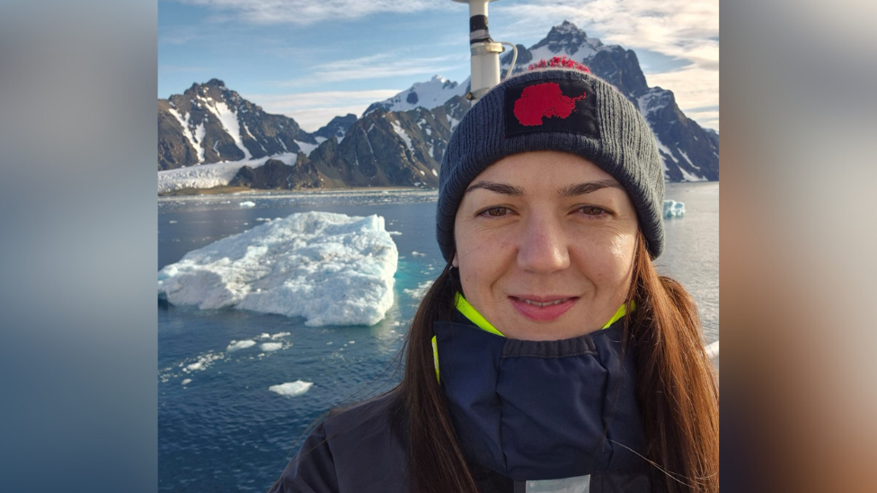 Capt Philippa Bowden has long brown hair and is wearing a large warm coat and woolly hat. Behind her is Antarctica, complete with snowy peaks and ice-laden seas.