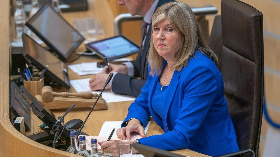 Alison Johnstone, wearing a blue suit, sits in the Presiding Officer's chair in the Scottish Parliament 