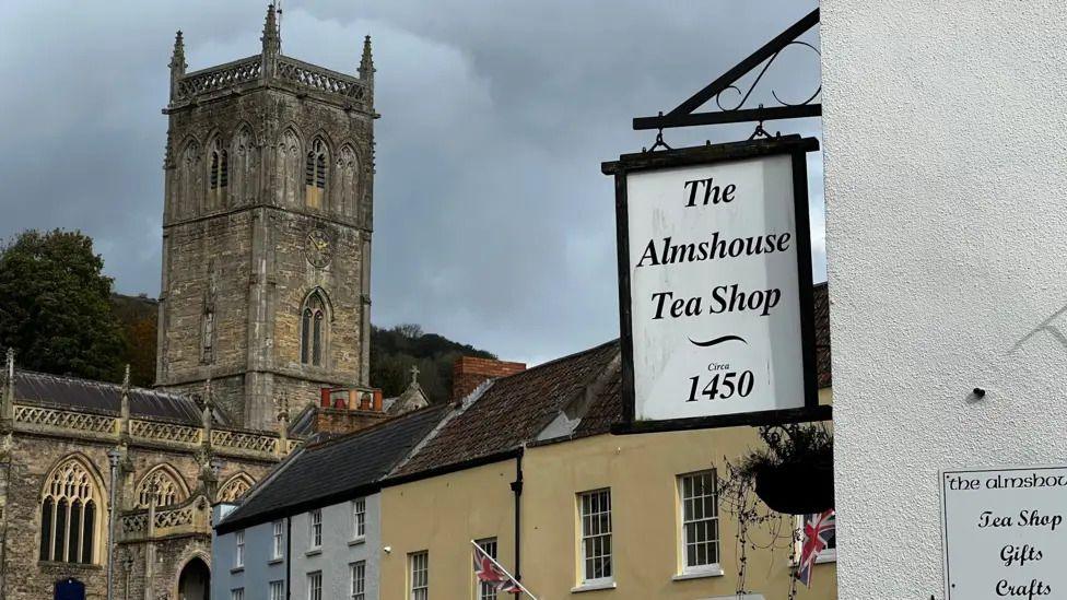 A view over Axbridge town square, towards the old church. In the foreground, a sign reads "The Alms House Tea Shop, established 1450".