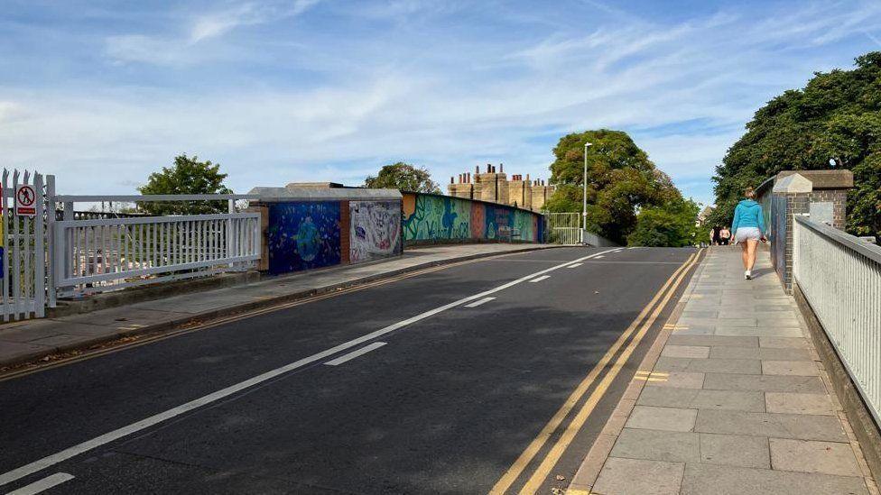 Mill Road bridge in Cambridge with a woman walking on the pavement. She is wearing shorts and a blue top. 