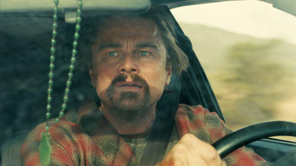Leonardo DiCaprio in One Battle After Another, driving a car and looking concerned while glancing in the rear-view mirror