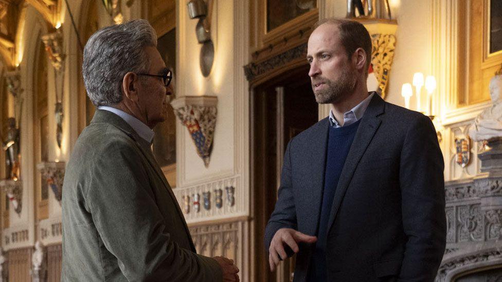 Prince William showing Eugene Levy inside Windsor Castle, they are in a large room or corridor with red carpet and ornate furnishings
