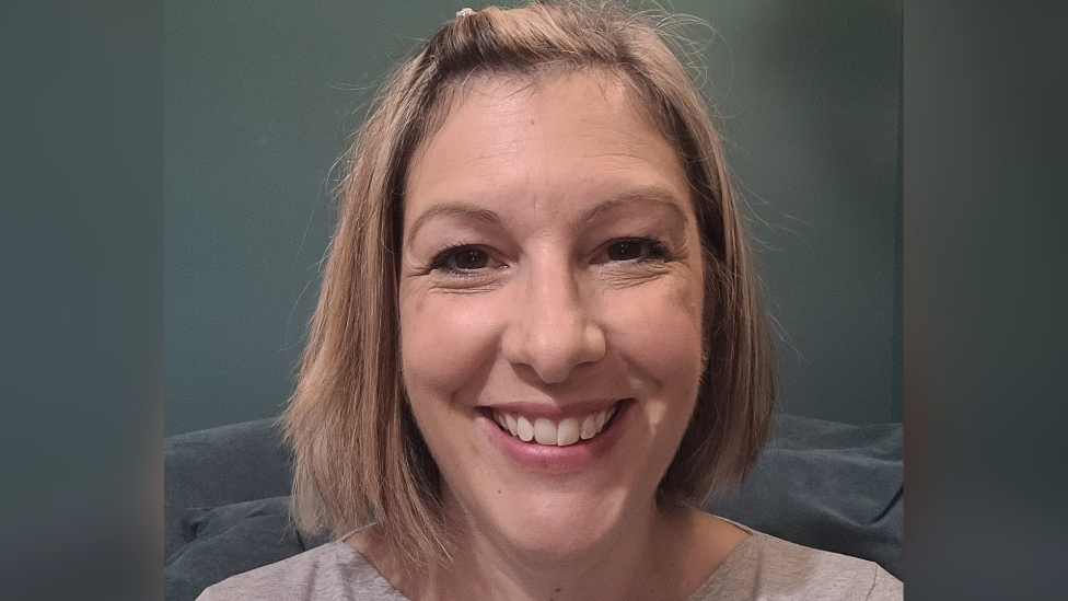 Zoe Bunter is smiling at the camera as she takes a photo of herself. She has blonde shoulder length hair and her fringe is clipped up to the side. She wears a light grey T-shirt and is stood against a plain dark grey background.