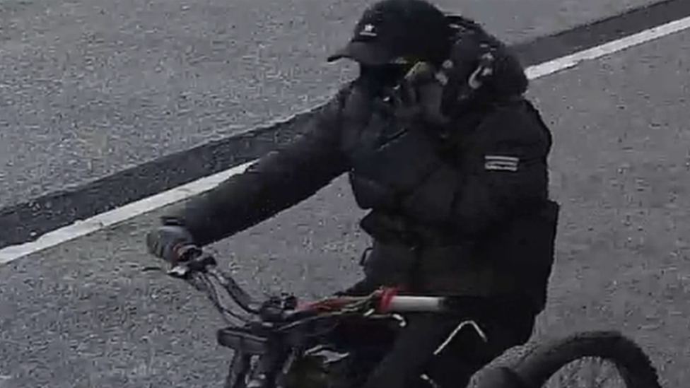 A still image of a man riding a red handled electric bike. He is dressed all in black with a bubble style coat, tracksuit bottoms, gloves, a baseball cap and a  covering over his lower face. His left hand is holding a mobile phone to his ear while his right hand is gripping the handle-bars of the bike. 