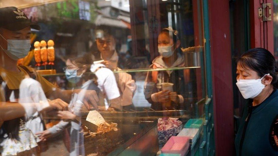 A woman wearing a face mask looks at a man cooking inside a restaurant in Beijing