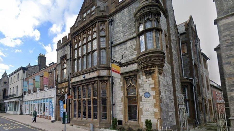 Google Street View of Dorset Museum - a gothic style stone building on the sloping High East Street in Dorchester. The large bay windows on the ground and first floors are made of lead-pane glass and framed with ornate stone.