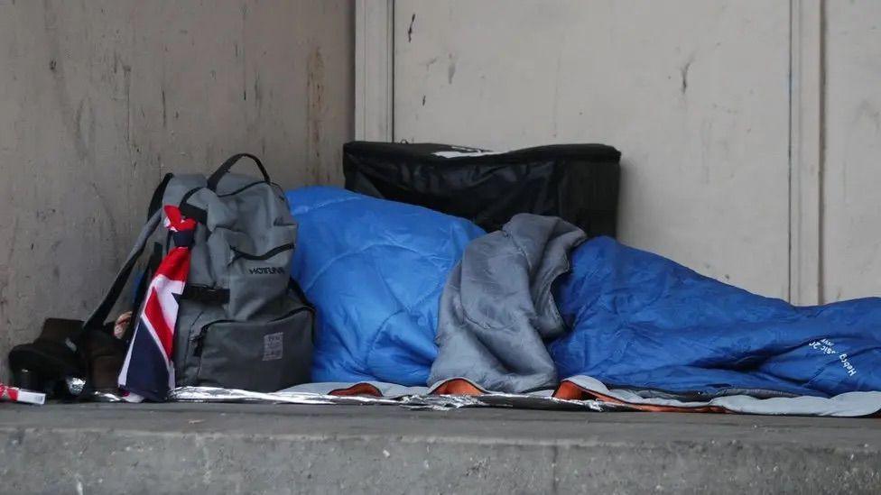 Someone in blue sleeping bag, rough sleeping, with grey backpack.