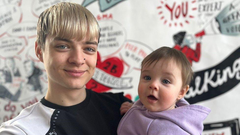 A photo of young dad Tyler holding his 10-month-old daughter Aurianna. Both are smiling. Tyler has dyed blonde hair and his long fringe is down to his eyebrows. He has a small black nose stud in and is wearing a black and white T-shirt. Aurianna has brunette hair and is wearing a purple jumper. It's a close up photo of their heads and shoulders and behind them is a mural on the wall with positive phrases on about being a dad.