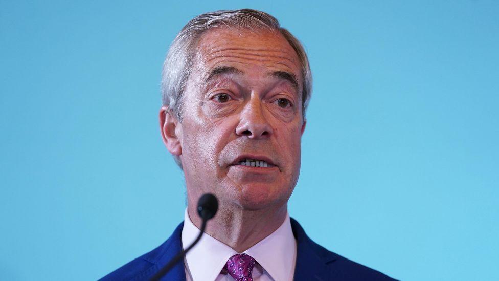 A man with grey hair, wearing a navy suit, purple shirt and purple tie, speaks into a microphone. The background is a light blue colour 