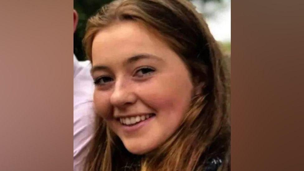 A close up image of Katie Simpson, a girl with long brown hair, smiling at the camera. 