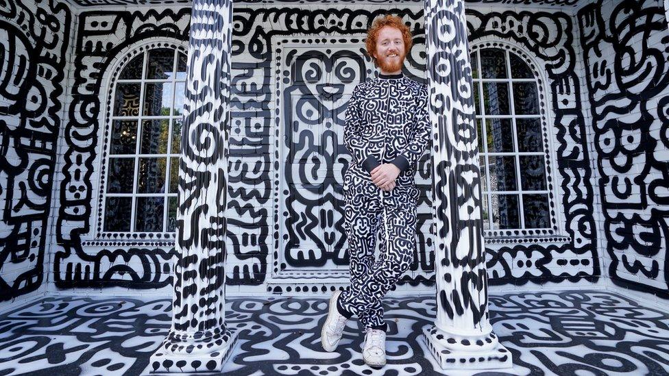 artist sam cox stands between two columns at the front of the house smiling while wearing a tracksuit covered in his doodles