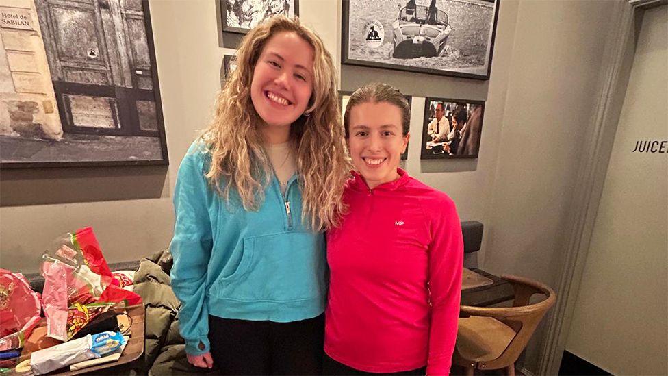 Megan, wearing a blue hoody and black leggings next to Harriet wearing a red top. They are in a room which has large framed photos in the background and a table next to them with sweets and biscuits.