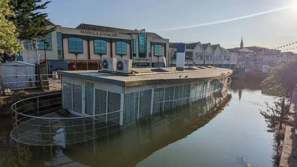 A boat which is half submerged in water. Shops behind it.