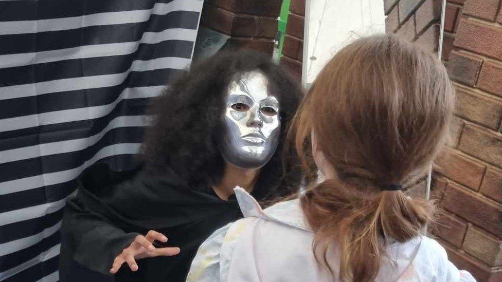 An actor in a silver mask with curly hair stares at a young girl, with hand extended in a claw shape, the girl with brown hair is seen here from the back of her head.