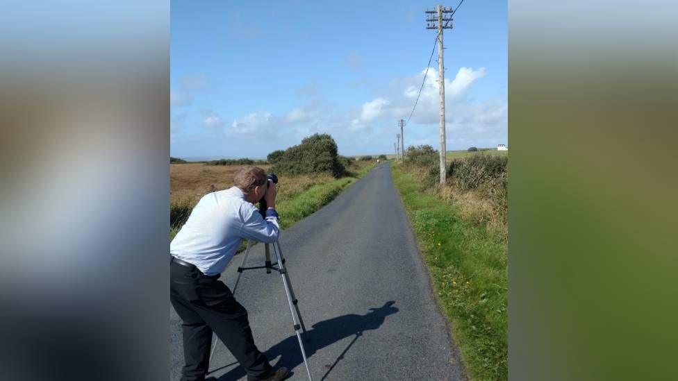 A man in a blue shirt takes a photograph of a line of telegraph poles running along the side of a tarmac road 