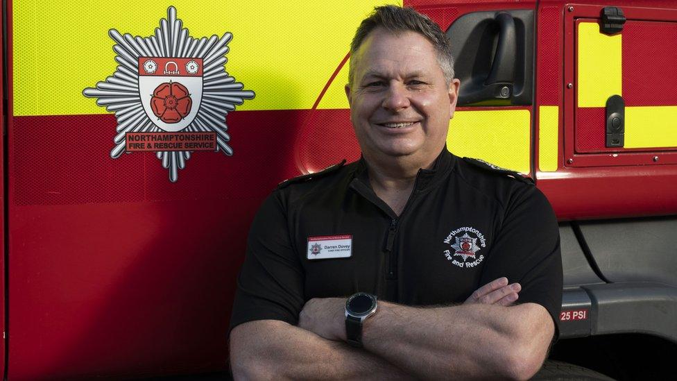 Man with short white hair wearing a blue and white striped shirt and standing in front of a fire engine