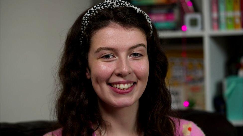 Elspeth in her apartment, sitting in front of a book case with pink lights