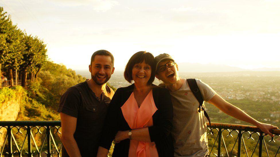 Anne Martinez with two young men, all smiling broadly, standing on a balcony overlooking a valley below dotted with trees and housing. Left to right: A man with short dark hair and a short dark beard, wearing a black T-shirt; Mrs Martinez with dark bobbed hair, wearing a dark jacket over an orange dress; a man wearing a light-coloured baseball cap, glasses and a light-coloured T-shirt.