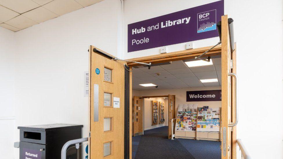 Pale wooden double doors stand open with a library beyond. A purple "hub and library" sign is above the door, and there is a "welcome" sign on the wall above a rack of leaflets.