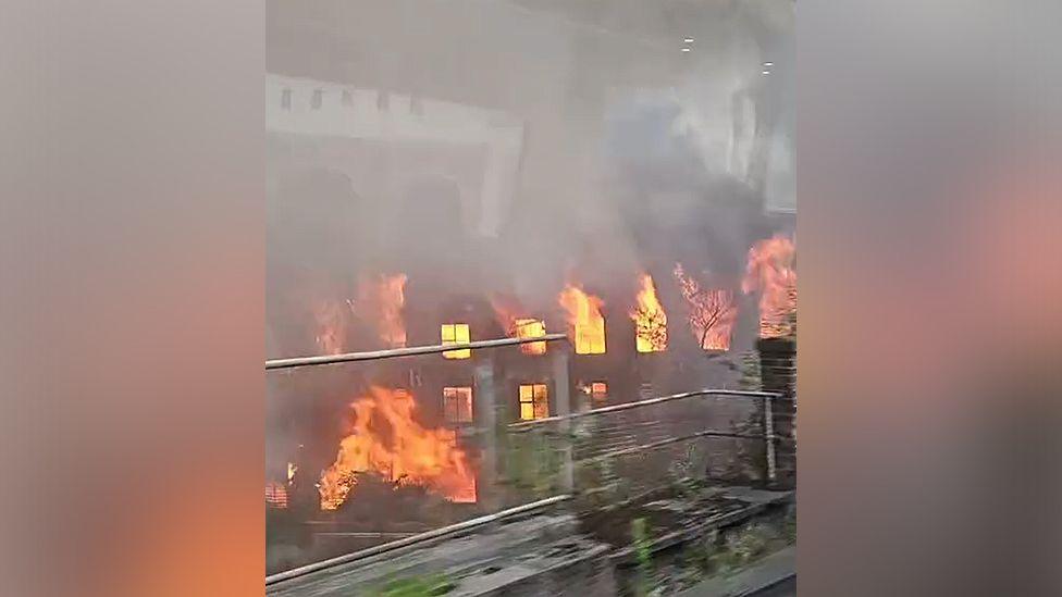Fire engulfs an old mill building in Manchester. Huge plumes of resulting smoke are visible across the city.