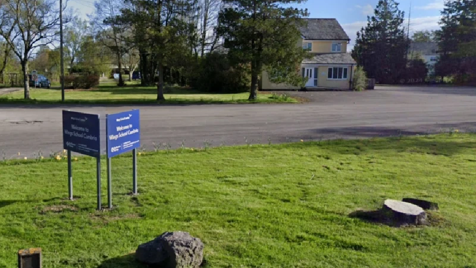 Blue signage on a small grass section reads 'Wings School Cumbria'. There is a large tarmaced driveway with trees and a large residential property.