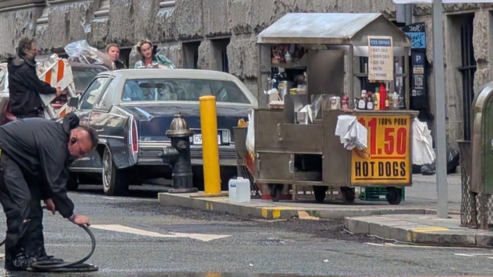 An American style Hot Dog stall and car are parked on a Liverpool city centre street. Film crew are moving cables and props.