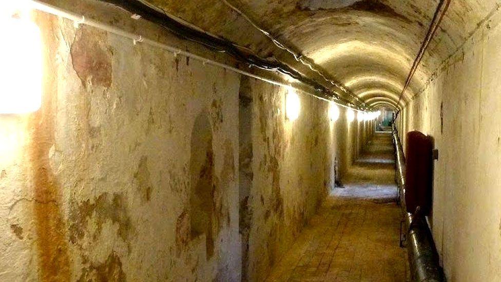 A tunnel beneath the Royal Pavilion in Brighton. The tunnel has straight side and a curved roof. The walls are white but the paint is peeling away. There is a square light on the left wall.