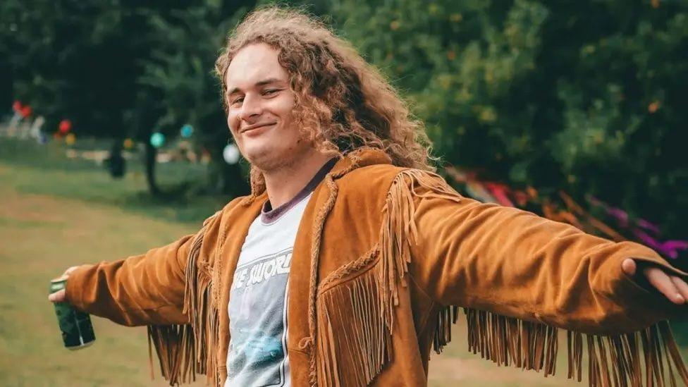A young man smiles at the camera as he holds his arms in a cross position. He has a drink in a can in one of his hands, and he is wearing a light brown jacket with tassels and a grey t-shirt. He has shoulder-length curly hair, which is light brown.