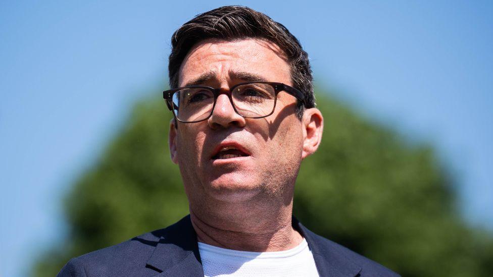 Andy Burnham, who has dark hair and glasses, speaks with the sun shining on his face, with a bright blue sky and tree in the background. He is wearing a navy suit jacket and white t-shirt. 