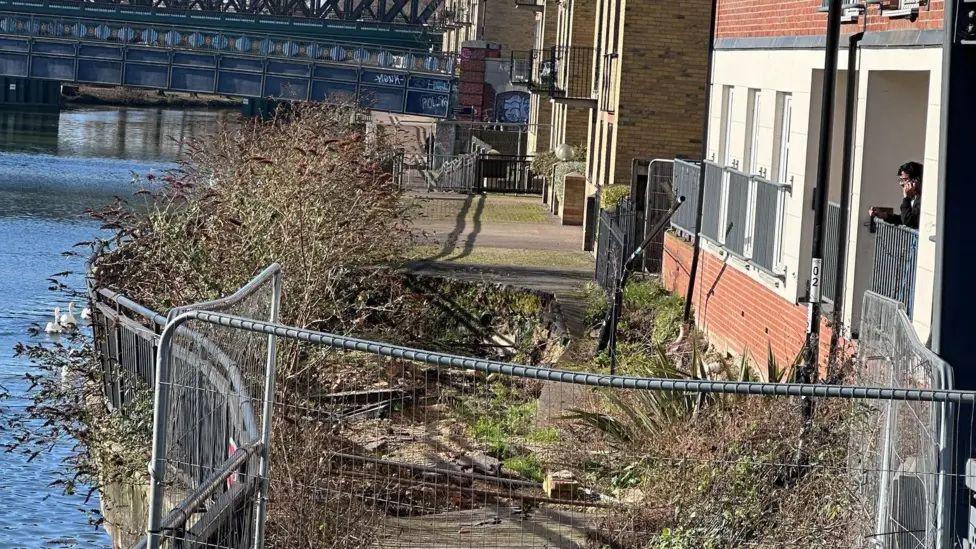A fenced off footpath with a sinkhole in it- placed between the River Nene and a block of flats on the other side of it.