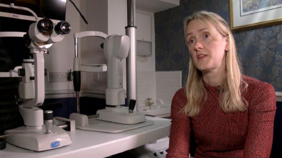 Claire Williams sat next to an eye test machine, wearing a red top and long blonde hair.