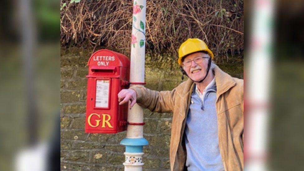 Reg Ling, wearing a blue top and light brown coat, leaning on the Frome Valentine's Lamp smiling. The lamp has colours painted on and is near a red post office.