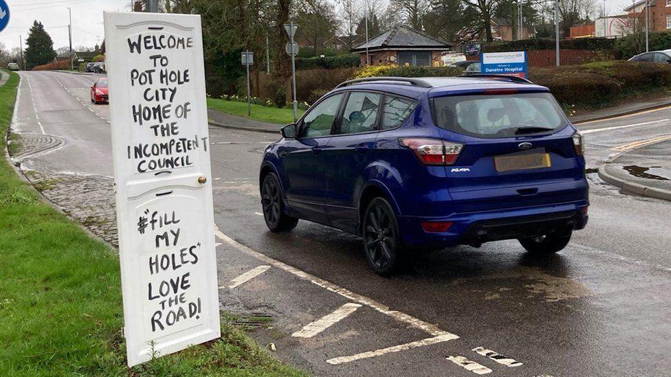 "Welcome to Pothole City" notice on a grass verge next to a small roundabout