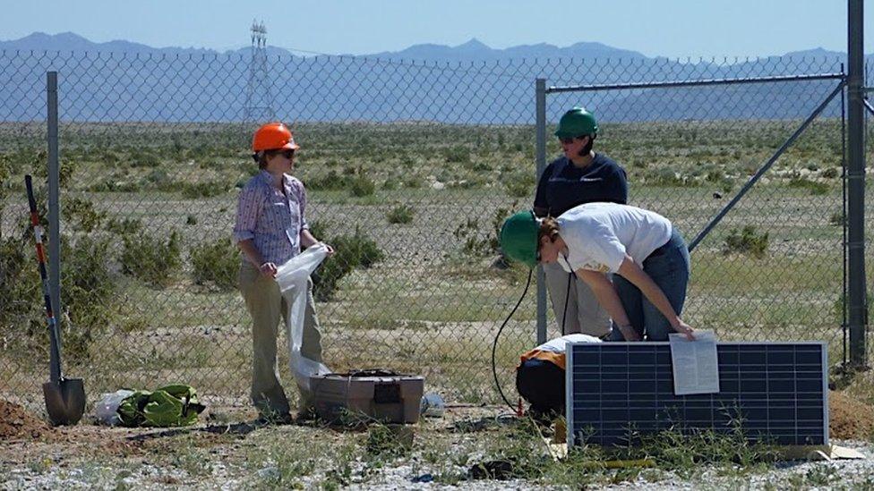 Three scientists handling sensor equipment in front of a chain link fence, desert behind