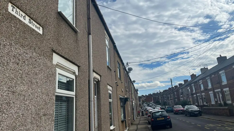 Terraced housing with the road sign saying Third Street. Cars are parked on both sides of the street.
