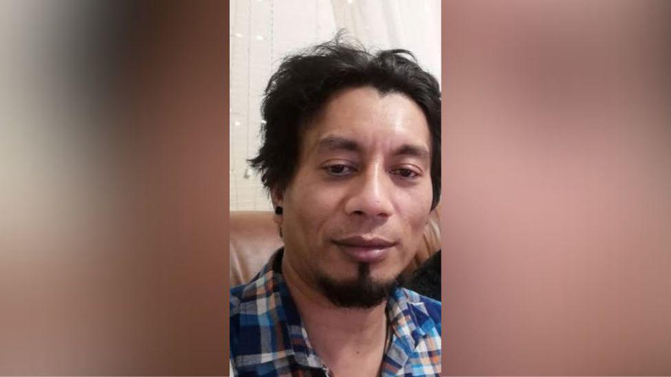 Ruben Alexandre Guimares who had short black hair and a black goatee. He is wearing a blue and orange gingham shirt and sitting on a brown sofa.