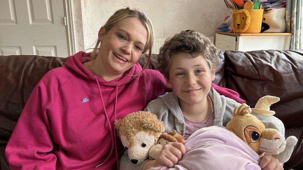 A mum in a pink hoddie and a young girl cuddle on the sofa