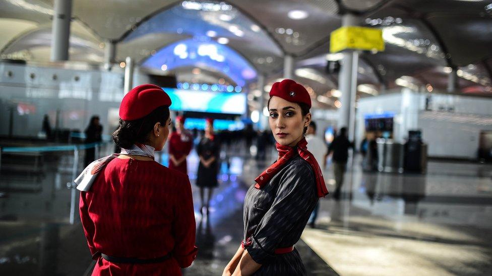 Two Turkish Airlines employees in distinctive red uniforms stand in the shining new halls of the airport, visible stretching behind them. One has her back to the camera, while the other has turned to look directly at the photographer