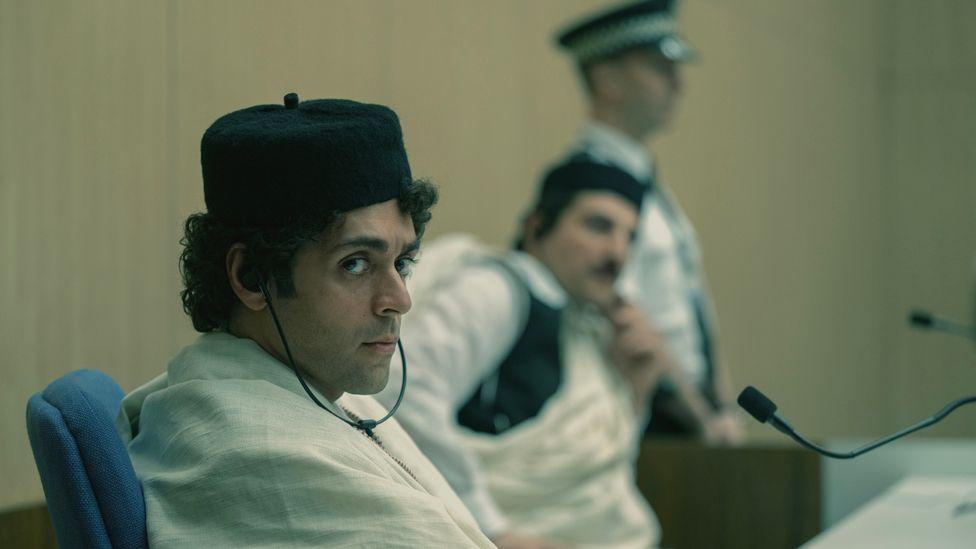 Ardalan Esmaili, playing Megrahi, is in the foreground sitting in court listening through headphones. Mudar Abbara, playing Fhimah, is sitting in the background, and a police officer is standing in the distance. 